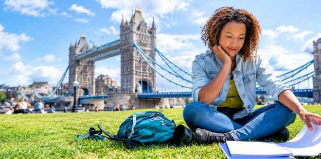 Young woman studying on a London lawn with books and a backpack, with Tower Bridge in the background.